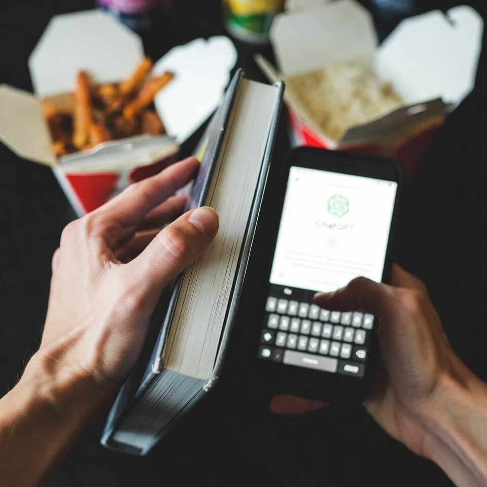 Hands holding old book and ChatGPT phone over takeout boxes