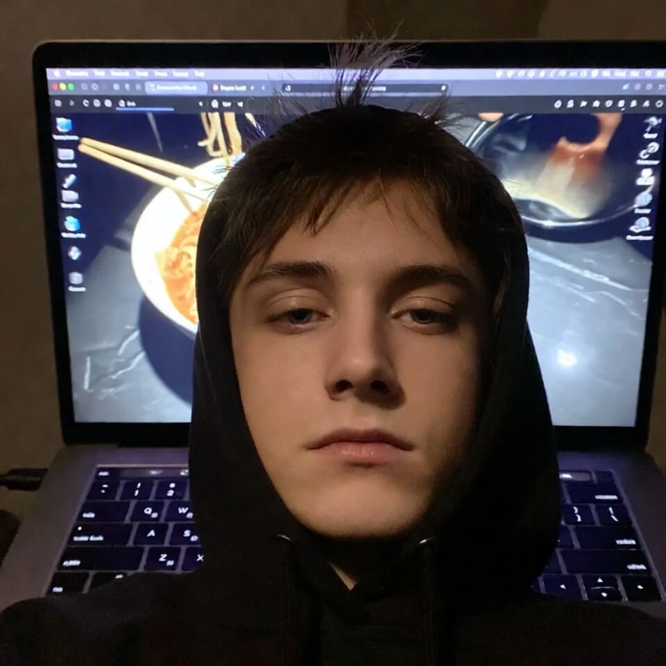 Exhausted student in hoodie, one eye closed, ramen reflected on laptop screen.